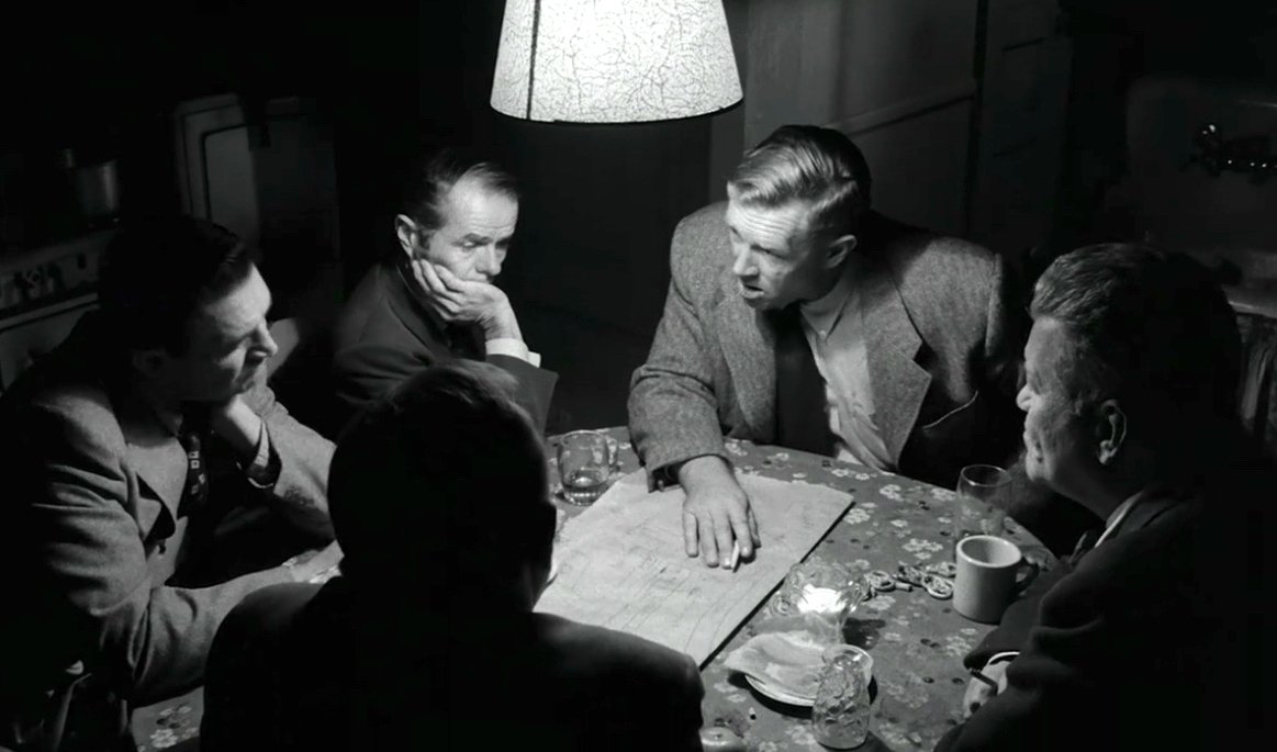 The conspirators preparing for the racetrack heist, huddled in a dark room beneath an overhead light.